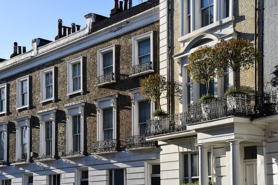 Photograph of a typical Chelsea terrace building with multiple floors and large sash windows. The building's facade features a mix of brickwork and decorative white stucco accents around the windows and doorway. Several small balconies with ornate black wrought iron railings extend from the upper floors, with potted plants and small trees placed on the balconies. The scene is illuminated by natural daylight, with clear blue skies in the background. The image captures the exterior of the property, illustrating an urban residential environment suitable for house removals or home relocation services. Man and a Van Chelsea often assist with packing, furniture transport, and loading processes, which are part of organized moves involving these types of historic terrace buildings.