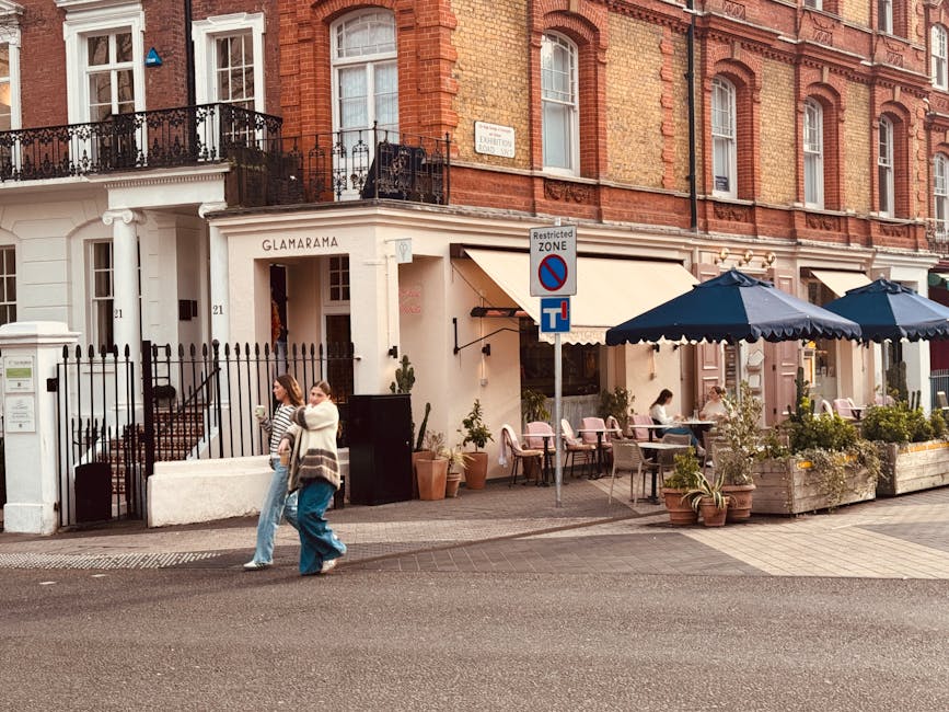 A street scene outside a building with a sign reading 'Glamarama' in Chelsea, featuring an outdoor seating area with pink chairs, potted plants, and large navy umbrellas. Two women walk past the entrance, one carrying a handbag while the other has a coat draped over her arm. The sidewalk is paved with concrete slabs, and a parking restriction sign is positioned near the outdoor seating. The building has a red brick exterior with white-framed windows and balconies with ornate black railings. Man and a Van Chelsea occasionally highlights house removals and furniture transport services, but in this image, the focus is on the exterior environment, which could relate to the logistics of home relocation or packing activities associated with moving services.