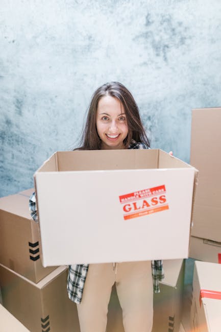A young woman with long brown hair smiling at the camera while holding an open cardboard box labeled 'FRAGILE GLASS' during a home relocation process inside a room with a light blue textured wall. She is dressed in casual clothing and is standing among other packed moving boxes of various sizes, some sealed with tape. Behind her, additional cardboard boxes are stacked, some with printed markings or labels, indicating organized packing. The scene captures a loading stage of furniture transport and packing for an eventual move, with evident focus on careful handling of fragile items as part of the professional services provided by Man and a Van Chelsea for house removals.