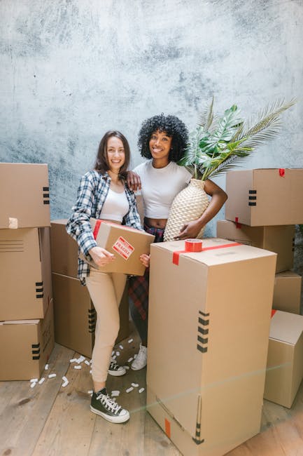 Two women engaged in a home relocation process indoors, surrounded by several cardboard boxes sealed with red tape, some placed on the floor and others stacked against a textured light grey wall. One woman, with light skin and long dark hair, is wearing a blue plaid shirt and beige pants, holding a cardboard box. The other woman, with dark skin and curly black hair, is dressed in a white T-shirt and patterned trousers, holding a woven pineapple-shaped decorative object with large green leaves. The scene depicts the loading or packing phase of furniture transport, with soft indoor lighting illuminating the space and a wooden floor beneath them. Some packing materials, such as white packing peanuts, are scattered on the floor. This image naturally aligns with the services of [COMPANY_NAME], a professional providing removals, packing, and home moving solutions, especially suited for house removals in areas like Chelsea, CHELSEA, as referenced in the page titled 'SW3 postcode removals tips for easy moves in Chelsea, CHELSEA' on [PAGE_URL].